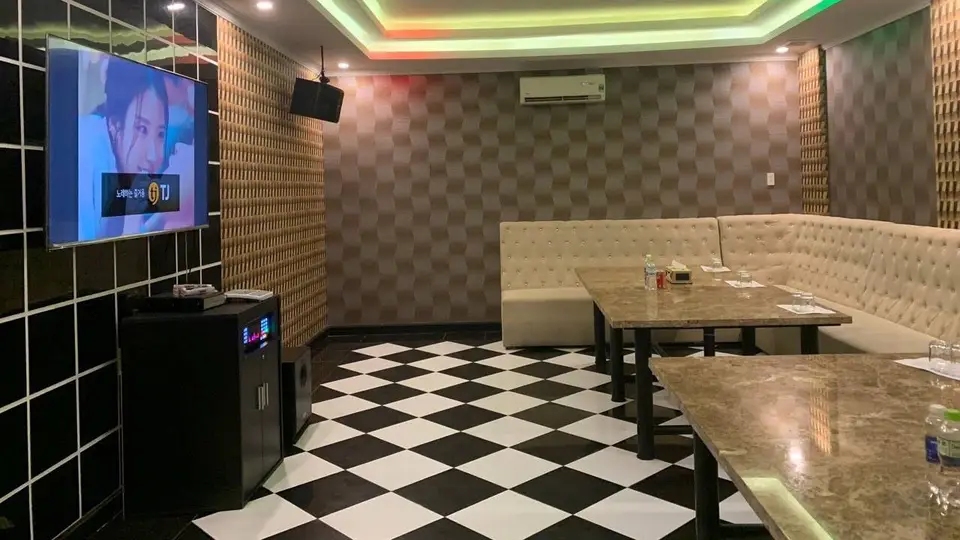 알록달록한 RGB LED 천장 조명과 TJ 노래방 기기, 크림색 소파 및 흑백 체크 바닥이 특징인 가라오케 룸