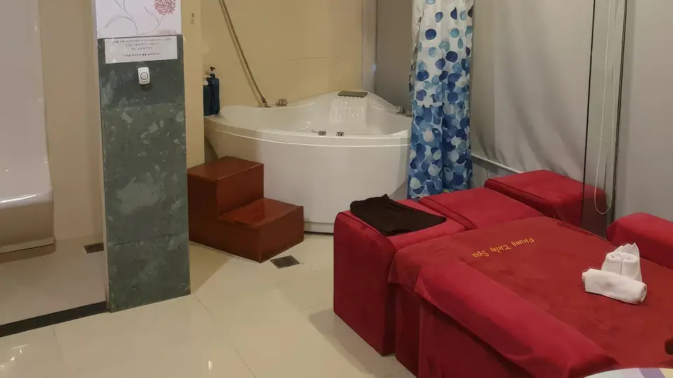 코너 자쿠지 욕조와 나란히 놓인 두 개의 붉은 마사지 베드, 대리석 기둥과 샤워 커튼이 어우러진 스파 룸