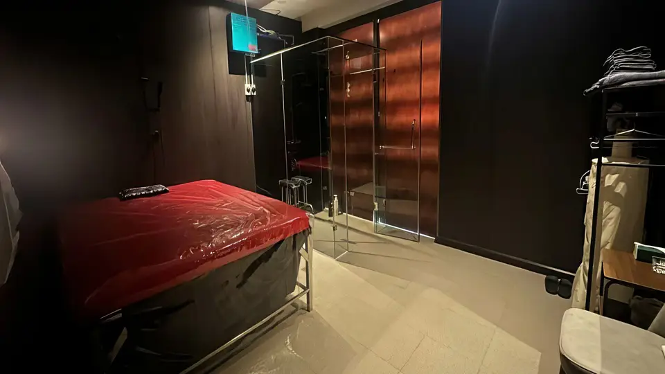 슈퍼맨 마사지 룸 내부 — 빨간 비닐 커버의 마사지 베드와 유리 파티션으로 구분된 샤워 공간, 어두운 우드 톤 벽면의 프라이빗 룸