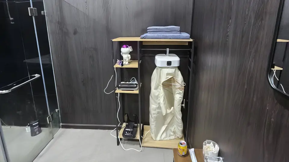 슈퍼맨 마사지 룸 내 어메니티 코너 — 접힌 타월과 가운이 비치된 오픈 행거 선반, 멀티탭 충전 공간, 소형 프로젝터 및 소품이 정돈된 수납 유닛